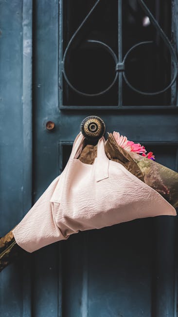 A person wearing brown gloves is holding a bouquet wrapped in soft pink paper with visible pink and green floral elements, likely roses or similar flowers, in front of a dark blue door. The bouquet is