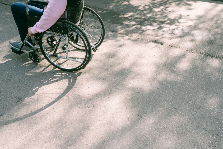 A person in a pink long-sleeve top and dark vest in a wheelchair is captured from behind, with their right arm resting on the armrest. The wheelchair has large black wheels and a metallic frame. The s