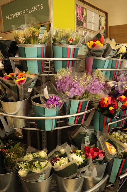 A display of various floral bouquets arranged in metal buckets on a multi-tiered stand inside a flower shop. The bouquets feature a range of colorful flowers including purple, yellow, pink, red, and w