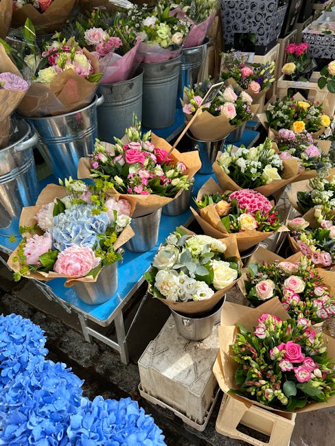 An array of fresh flower bouquets arranged on a table at a floral shop, featuring pastel pink and white roses, blue hydrangeas, and greenery, wrapped in brown craft paper and placed in metallic bucket