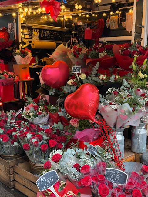 A vibrant display of fresh flowers and floral gifts at a flower stall, featuring predominantly red roses, white baby's breath, and greenery arranged in bouquets and wrapped in tissue paper and clear w